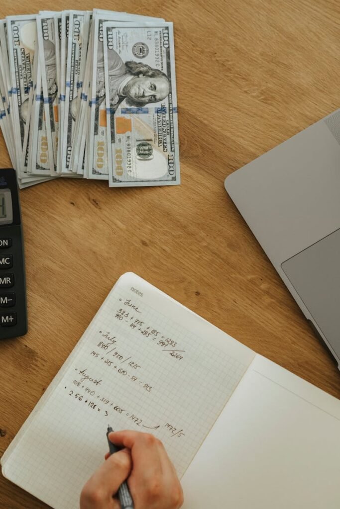 Hands using calculator and notebook, planning finances, with cash and laptop on wooden table.